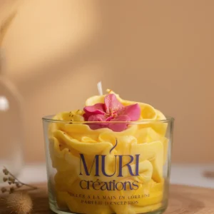 Bougie artisanale Douceur des îles de la marque Muri Créations, présentée dans un verre avec une cire jaune texturée effet chantilly et décorée d'une fleur rose. Bougie parfumée au monoï, noix de coco et vanille, coulée à la main en Lorraine.