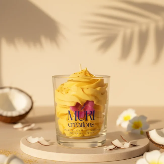 Bougie artisanale Douceur des îles de Muri Créations à la cire fouettée jaune, parfumée au monoï, noix de coco et vanille, mise en scène avec des fleurs de frangipanier et des copeaux de coco.
