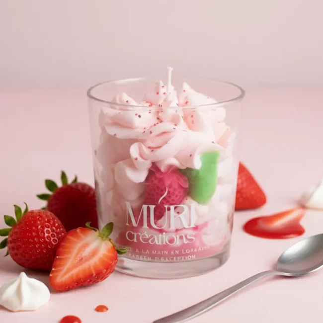 Bougie parfumée gourmande à la fraise de la marque artisanale Muri Créations, imitant un dessert avec de la cire fouettée rose effet chantilly dans un verre, entourée de fraises fraîches, de meringues et d'une cuillère sur fond rose.
