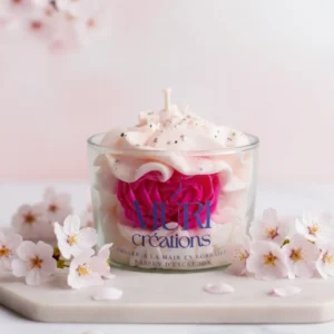 Bougie parfumée artisanale fleur de cerisier par Muri Créations, coulée à la main en Lorraine. Design gourmand en cire fouettée avec rose fuchsia incrustée, décorée de paillettes et entourée de fleurs de sakura sur fond rose.