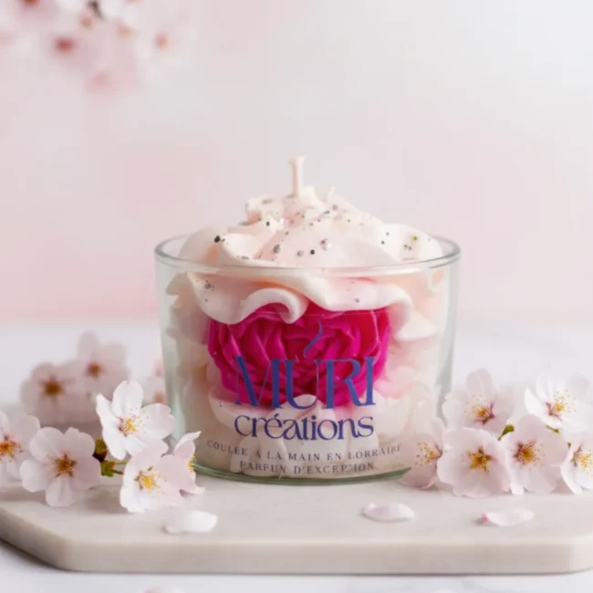 À propos 7 Bougie parfumée artisanale fleur de cerisier par Muri Créations, coulée à la main en Lorraine. Design gourmand en cire fouettée avec rose fuchsia incrustée, décorée de paillettes et entourée de fleurs de sakura sur fond rose.