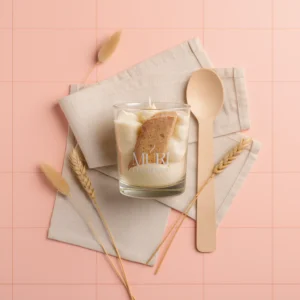 Bougie gourmande artisanale Muri Créations parfumée à la brioche, présentée dans un verre avec une texture crémeuse et un décor de biscuit. Mise en scène esthétique sur fond rose carrelé avec une cuillère en bois, une serviette en lin beige et des épis de blé séchés.