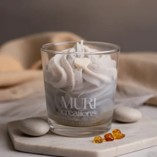 Bougie parfumée artisanale à l'ambre gris de la marque Muri Créations, présentée dans un verre avec une cire fouettée grise effet chantilly décorée d'un cœur, posée sur un support en pierre entouré de cristaux d'ambre.