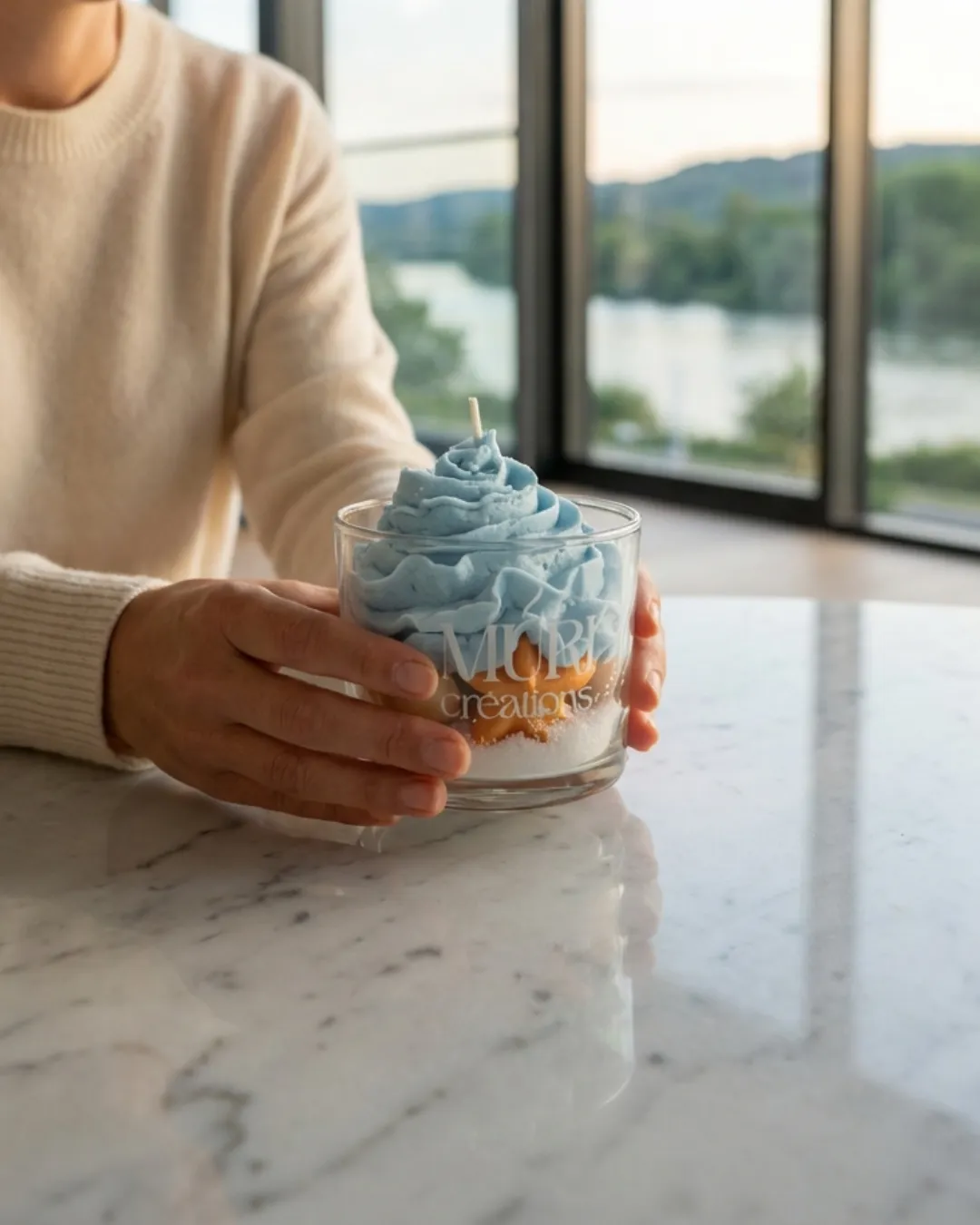 Bougie 150ml - BRISE MARINE 3 Mains tenant la bougie artisanale Brise Marine de la collection printemps été 2026 de la marque Muri Créations. Cette bougie décorative en verre présente une cire fouettée bleue rappelant l'écume et des étoiles de mer, posée sur une table en marbre devant une fenêtre.