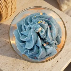 Vue de dessus de la bougie artisanale Brise Marine en cire fouettée bleue ornée de paillettes, issue de la collection printemps-été 2026 de la marque Muri Créations, posée sur une surface en pierre naturelle.