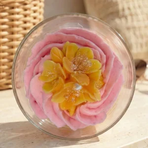 Vue de dessus d'une bougie artisanale au parfum floral de la collection printemps été 2026 par Muri Créations, présentant une cire fouettée rose et une fleur jaune pailletée au centre dans un contenant en verre.