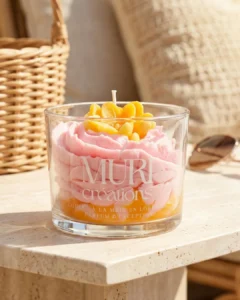 Accueil 3 Bougie artisanale gourmande Muri Créations au parfum floral, issue de la collection printemps été 2026. Bougie en verre avec cire rose effet crème fouettée sur base orange et décorations florales jaunes, coulée à la main en Lorraine, posée dans une ambiance estivale.
