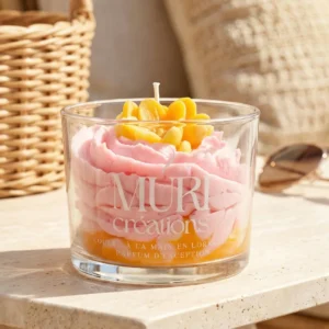 Bougie artisanale gourmande Muri Créations au parfum floral, issue de la collection printemps été 2026. Bougie en verre avec cire rose effet crème fouettée sur base orange et décorations florales jaunes, coulée à la main en Lorraine, posée dans une ambiance estivale.