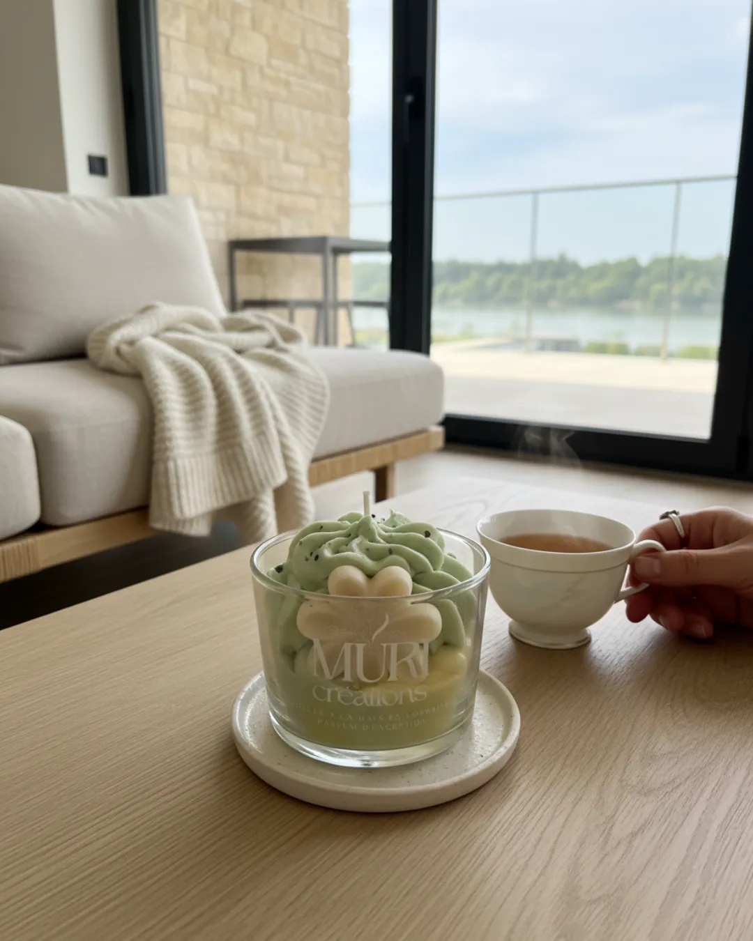 Bougie 150ml - JASMIN 3 Bougie artisanale au parfum floral de jasmin de la marque Muri Créations, dotée d'un design gourmand effet chantilly vert et fleur, posée sur une table basse en bois à cÎté d'une tasse de thé. Ambiance cocooning face à une baie vitrée pour présenter la collection printemps été 2026.
