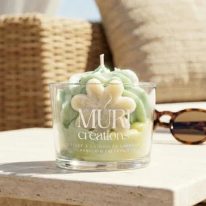 Bougie artisanale au parfum floral de jasmin de la marque Muri Créations, collection printemps été 2026. Gros plan sur une bougie gourmande en verre avec cire fouettée vert pastel et insert fleur, coulée à la main en Lorraine, posée sur une table en pierre au soleil.