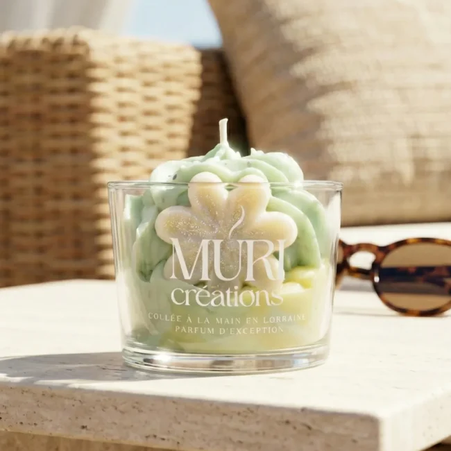 Bougie artisanale au parfum floral de jasmin de la marque Muri Créations, collection printemps été 2026. Gros plan sur une bougie gourmande en verre avec cire fouettée vert pastel et insert fleur, coulée à la main en Lorraine, posée sur une table en pierre au soleil.