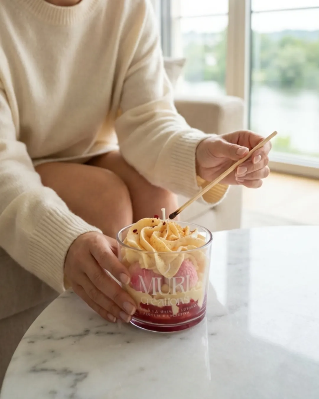 Bougie 150ml - LITCHI 3 Femme allumant la mèche d'une bougie artisanale parfum Litchi de la marque Muri Créations, issue de la collection printemps été 2026, au design gourmand évoquant un dessert glacé sur une table en marbre.