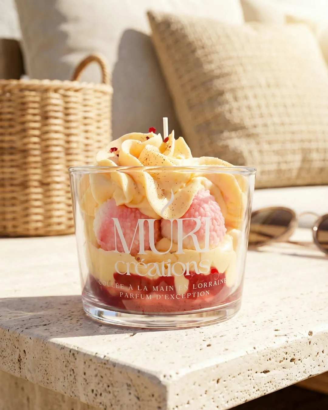 Bougie 150ml - LITCHI 1 Bougie artisanale gourmande parfum Litchi de la marque Muri Créations, collection printemps été 2026. Cette création coulée à la main en Lorraine imite un dessert avec sa cire fouettée et ses sphères roses dans un verre transparent, posée sur une table en pierre ensoleillée.
