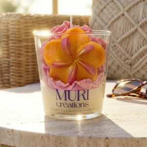 Bougie artisanale gourmande au parfum monoï passion de la marque Muri Créations, collection printemps été 2026. Cette bougie en verre, coulée à la main en Lorraine, est décorée d'une cire fouettée rose et d'une fleur tropicale orange pailletée, mise en scène sur une table en pierre dans une ambiance estivale ensoleillée.