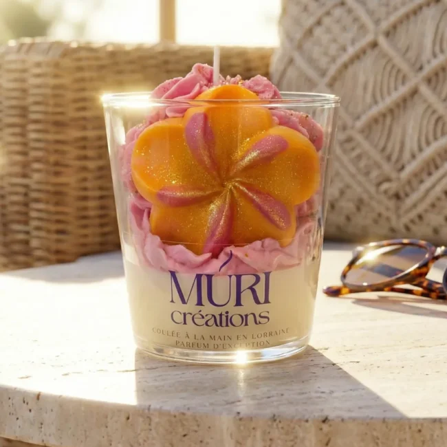 À propos 10 Bougie artisanale gourmande au parfum monoï passion de la marque Muri Créations, collection printemps été 2026. Cette bougie en verre, coulée à la main en Lorraine, est décorée d'une cire fouettée rose et d'une fleur tropicale orange pailletée, mise en scène sur une table en pierre dans une ambiance estivale ensoleillée.