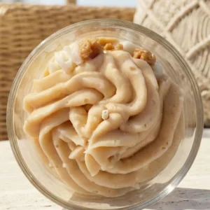 Vue de dessus de la bougie artisanale parfum popcorn de la marque Muri Créations. Cette bougie gourmande présente une cire fouettée beige texturée style chantilly ornée d'éclats de popcorn, issue de la collection printemps été 2026, avec un arrière-plan flou en osier et macramé.