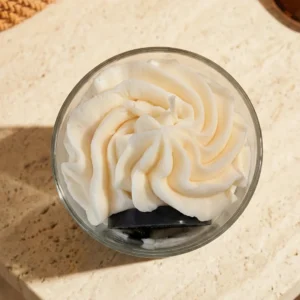 Vue de dessus d'une bougie artisanale gourmande au parfum réglisse signée Muri Créations. Cette bougie de la collection printemps été 2026 présente une cire fouettée blanche style chantilly sur une base sombre, posée sur une surface en travertin.