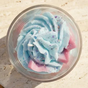 Vue de dessus de la bougie artisanale Muri Créations au parfum bonbon bouteille : cire fouettée bleue et rose style chantilly parsemée de paillettes et de fondants, collection printemps été 2026.
